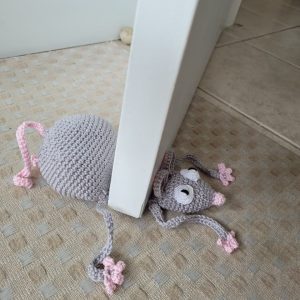 Peso de porta em crochê em formato de ratinho cinza com dedinhos rosa, apoiado no rodapé de uma porta branca.