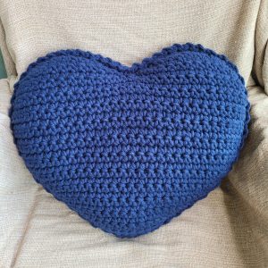 Almofada de crochê em formato de coração, cor azul-marinho, sobre poltrona de linho claro.