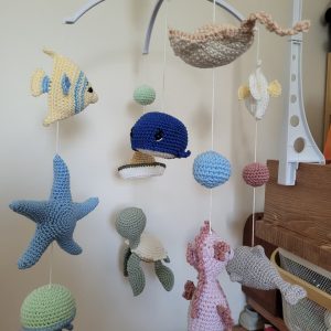 Móbile de berço em crochê com tema fundo do mar, peixe, baleia azul, estrela-do-mar, tartaruga e cavalo-marinho pendurados numa haste branca.
