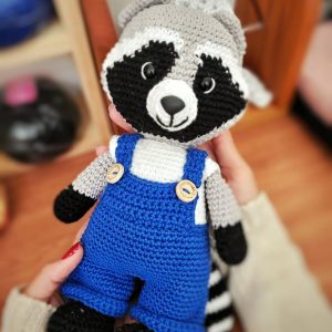 Amigurumi de guaxinim em crochê cinza e preto, vestindo jardineira azul com botões de madeira, segurado na mão.