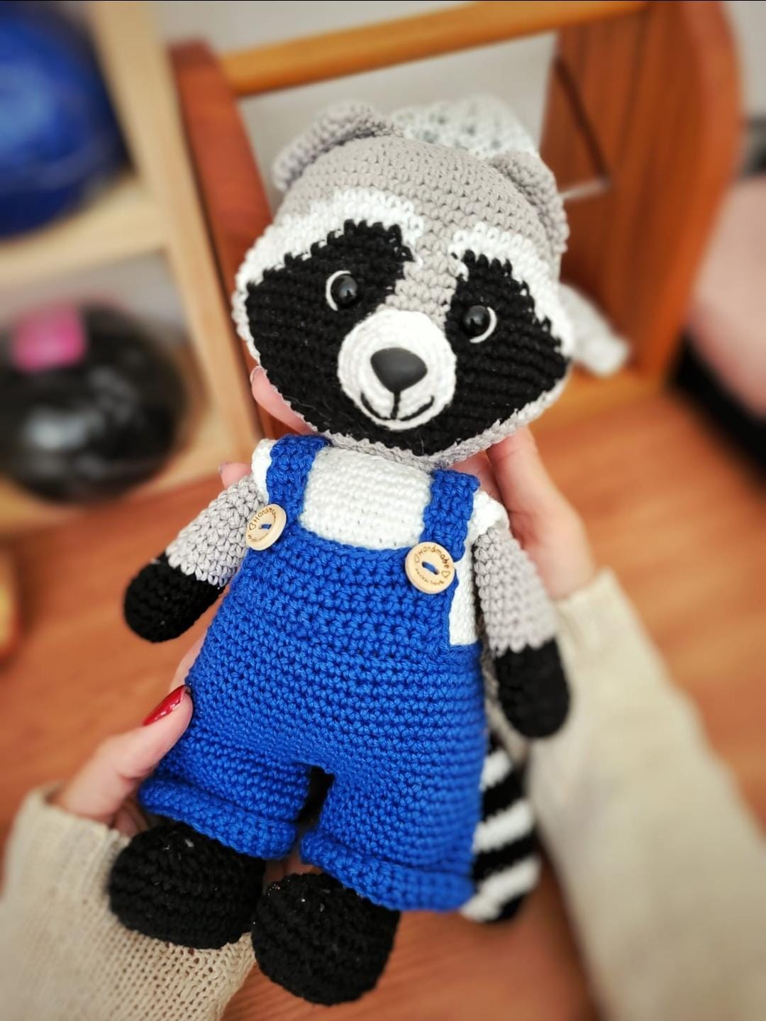 Amigurumi de guaxinim em crochê cinza e preto, vestindo jardineira azul com botões de madeira, segurado na mão.