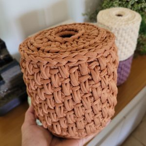 Capa de crochê terracota para rolo de papel higiênico, segurada na mão, mostrando a textura trançada.