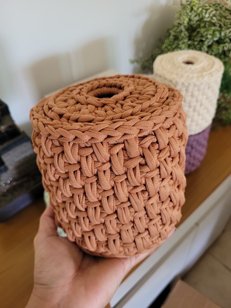 Capa de crochê terracota para rolo de papel higiênico, segurada na mão, mostrando a textura trançada.