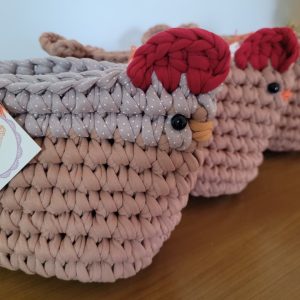 Três cestos de crochê em formato de galinha, cor terracota, com cristas vermelhas e olhos pretos, um deles com a tag Café Crochet pendurada.