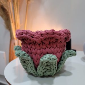 Cachepot de crochê em formato de flor, corpo rosé e pétalas verde-sálvia, vazio, sobre mesa branca com folhagem seca ao fundo.