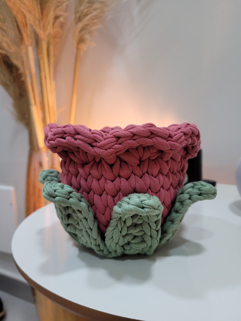Cachepot de crochê em formato de flor, corpo rosé e pétalas verde-sálvia, vazio, sobre mesa branca com folhagem seca ao fundo.