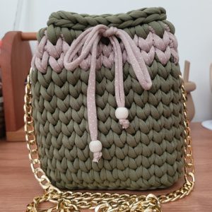 Bolsa balde em crochê bege com faixa poá rosé, alça de corrente dourada, sobre mesa de madeira.