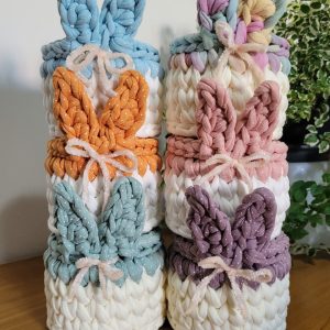 Seis cestinhas de crochê com orelhas de coelho em cores diferentes (azul, laranja poá, verde-água poá, multicolor pastel, rosa poá e roxo poá), empilhadas em duas torres.