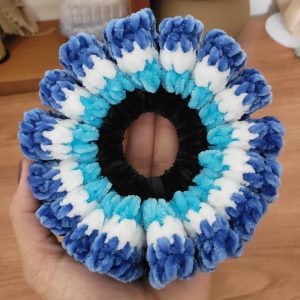 Scrunchie de crochê em pelúcia azul, branco e turquesa, segurado na palma da mão.