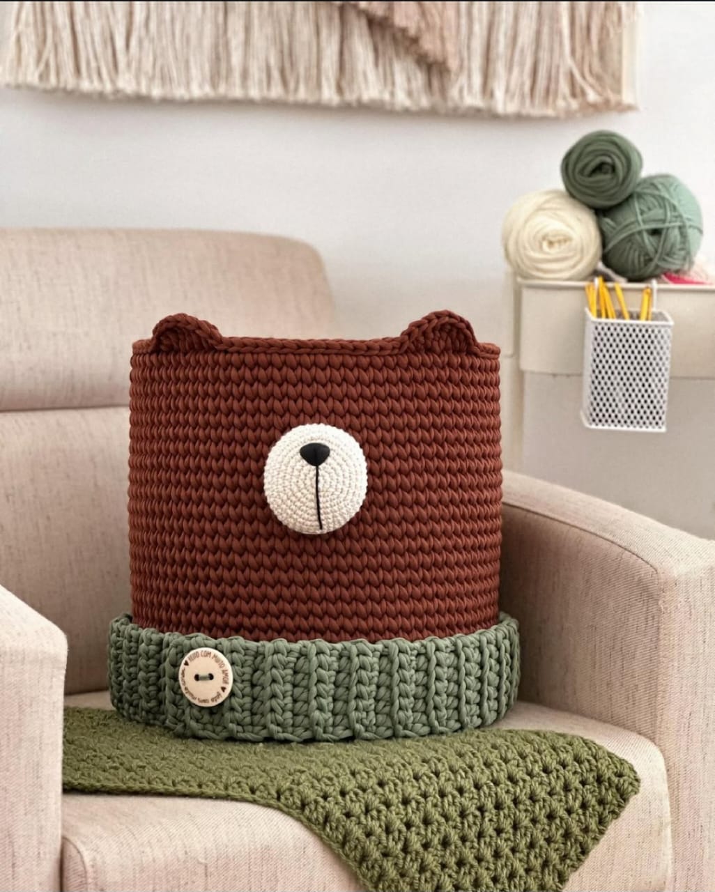 Cesto organizador de crochê em formato de urso terracota, focinho branco, faixa verde-sálvia com botão de madeira, alças de couro.