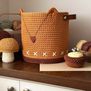 Cesto organizador de crochê caramelo com base marrom e detalhes em ponto-cruz creme, alça de couro, ao lado de cogumelos de crochê decorativos.