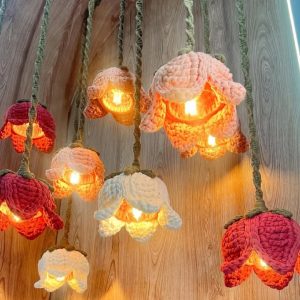 Várias luminárias de crochê em formato de flor, penduradas, em tons de rosa, bordô e cru, acesas, sobre fundo de madeira.
