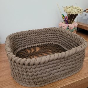 Cesto organizador oval de crochê cor taupe, com bandeja de madeira vazada em padrão de folhas no fundo.