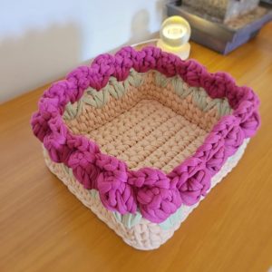 Cesto retangular de crochê cru com borda de flores em relevo magenta e folhas verde-sálvia.