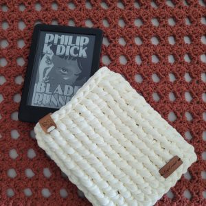 Capa de crochê cru para Kindle, com o dispositivo real (capa do livro Blade Runner na tela) ao lado, sobre tapete de crochê terracota.