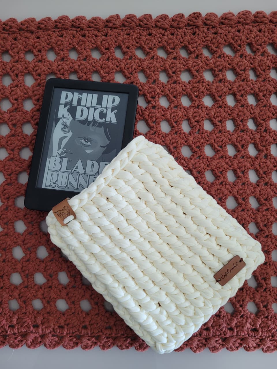 Capa de crochê cru para Kindle, com o dispositivo real (capa do livro Blade Runner na tela) ao lado, sobre tapete de crochê terracota.