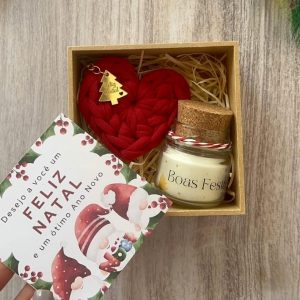 Box de Natal aberto com chaveiro coração de crochê vermelho, charme dourado de arvorezinha, e mini vela Boas Festas, sobre mesa de madeira.