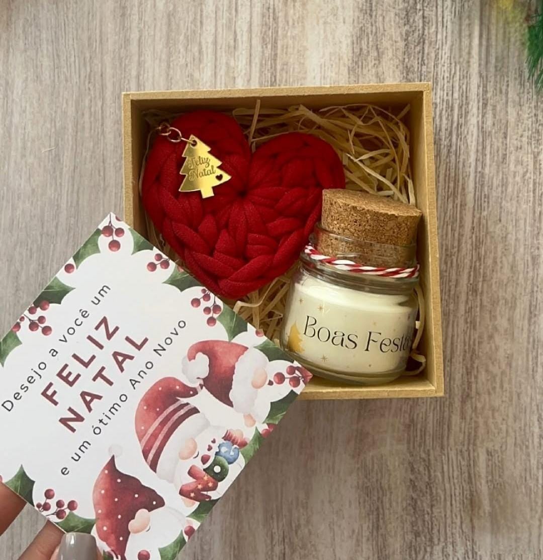 Box de Natal aberto com chaveiro coração de crochê vermelho, charme dourado de arvorezinha, e mini vela Boas Festas, sobre mesa de madeira.