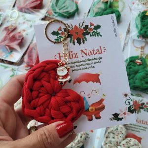 Chaveiro coração de crochê verde com pingente esmaltado de pinguim, preso a cartão Feliz Natal, segurado na mão.
