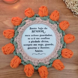 Mandala de crochê com moldura de tulipas laranja e folhas verdes, Oração do Anjo da Guarda impressa no centro, sobre mesa de madeira.