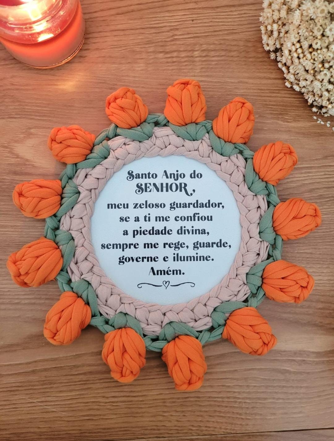Mandala de crochê com moldura de tulipas laranja e folhas verdes, Oração do Anjo da Guarda impressa no centro, sobre mesa de madeira.
