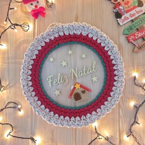 Guirlanda de crochê vermelha e branca com poá natalino, Feliz Natal escrito no centro e casinha decorativa, pendurada com luzinhas ao redor.