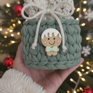 Saquinho de crochê verde-sálvia com cordão cru e charme de biscoito de gengibre, em frente a árvore de Natal decorada.