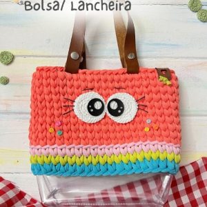 Bolsa lancheira de crochê coral com carinha de olhos grandes aplicados, faixa colorida na base e alças de couro, sobre fundo xadrez vermelho.