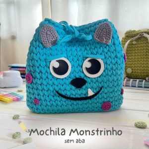Mochila de crochê turquesa com carinha de monstrinho de dois olhos, orelhas cinza e detalhes rosa, sobre mesa clara.