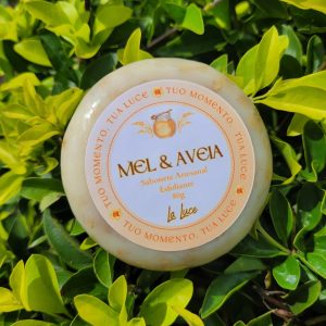 Sabonete artesanal La Luce Mel & Aveia, 80g, rótulo pêssego com ícone de pote de mel, sobre folhagem verde.