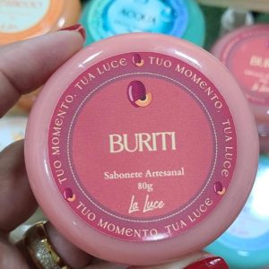 Sabonete artesanal La Luce Buriti, 80g, rótulo coral com ilustração de fruta buriti, segurado na mão.