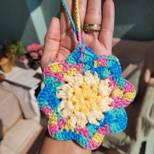 Bolsinha de crochê em formato de flor, tons pastel multicoloridos com centro creme e amarelo, pendurada na mão.