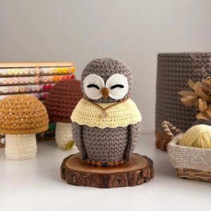 Corujinha de crochê cinza com olhos fechados brancos e capa amarela com pingente, sobre fatia de madeira entre cogumelos decorativos.